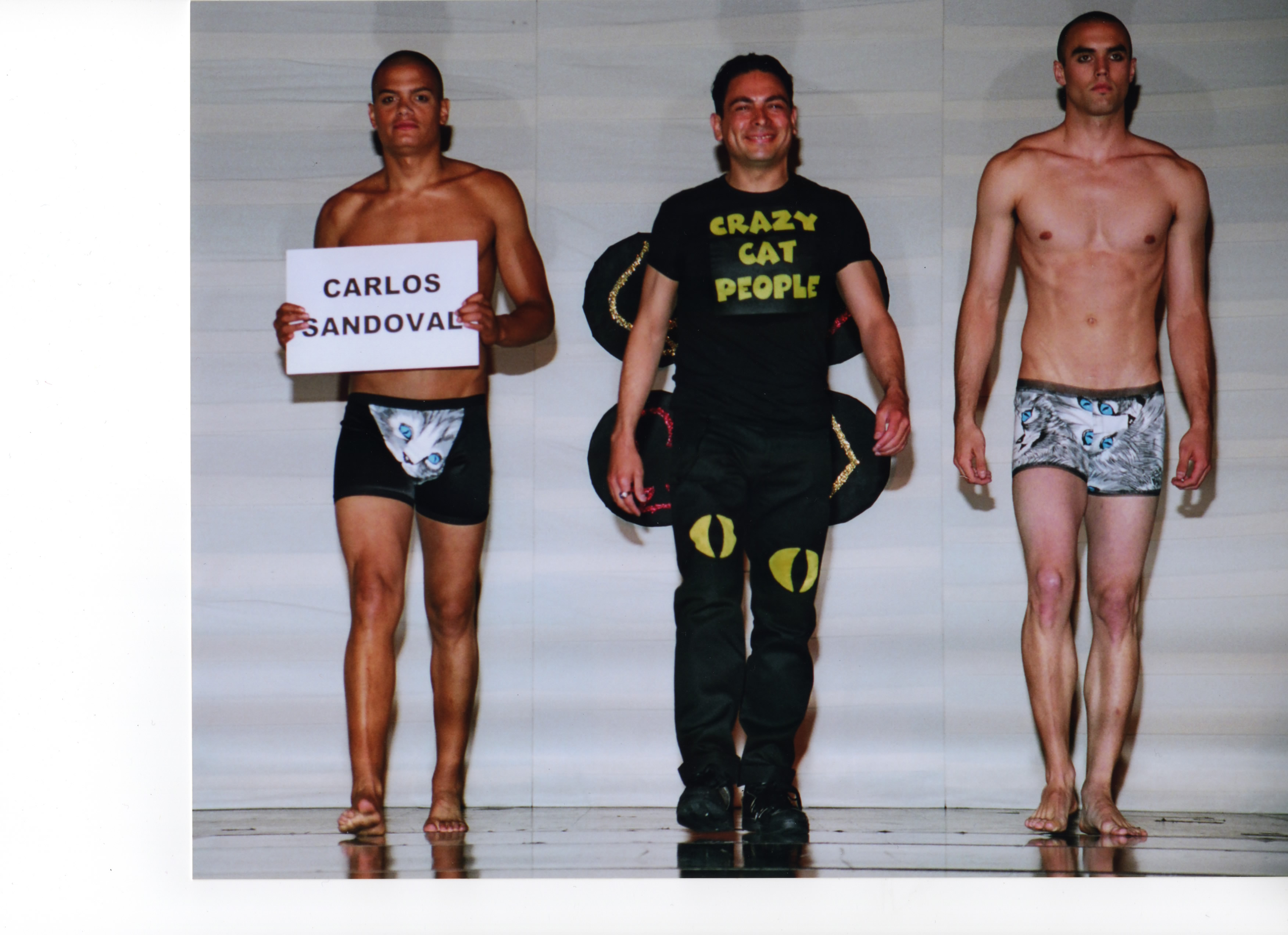 Fashion designer Carlos Sandoval wearing a Crazy Cat People shirt with cat eyes on the pant legs. On each side is a male model wearing cat-themed swimwear 